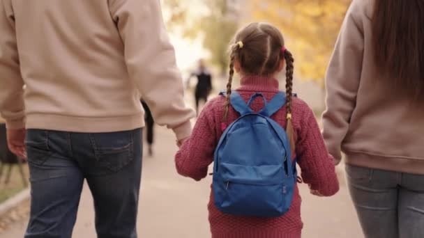 Anne ve baba küçük bir kızı sırtında sırt çantasıyla tutarlar, mutlu bir aile, sonbaharda okul bahçesinde yürürler, anne, baba ve çocuk omuzlarının üzerinde çanta taşırlar, birinci sınıftaki çocuğa eşlik ederler. — Stok video