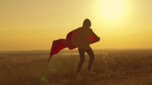 Glad tonåring flicka kör spela superhjälte vid solnedgången, barn på ett grönt fält i en röd regnrock leksak i strålarna av ljus, barns drömspel, kid drömmar om att bli en superhjälte i bländning av solen — Stockvideo