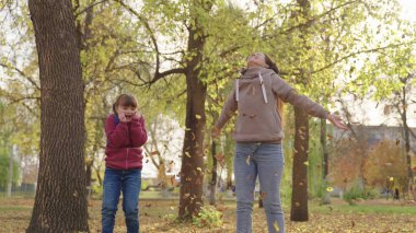 Okul çantalı küçük kız annesiyle sarı yapraklar fırlatıyor, çocuklar sonbahar oyunları hayal ediyor, mutlu aile, çocuk okuldan sonra okul bahçesinde annesiyle oynuyor, neşeli çocuk altın yaprak döküyor.