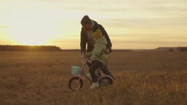 Küçük çocuk gün batımında babasıyla bisiklet sürmeyi öğreniyor, mutlu aile, çocukluk hayali bisiklet sürmeyi öğreniyor, çocuk pedalları güneş altında, kız ve baba oyun oynarken gülüyor, babalar günü