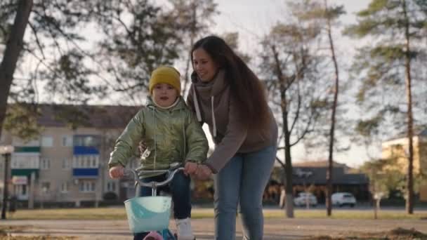 Mother teaches little child to ride bike in the city, kid rides on sidewalk, happy family, baby plays and laughs with mom, learn ride, make childhood dream come true girls, daughter and mom are funny — Stock Video