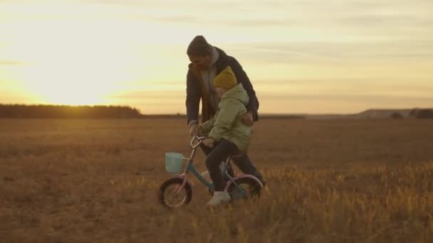 Маленький ребенок учится ездить на велосипеде с отцом на закате, счастливая семья, детская мечта научиться ездить на велосипеде, педали на солнце, девочка и папа смеются весело во время игры, День отца — стоковое видео