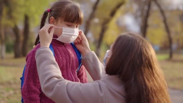 Mutter setzt Kleinkind eine medizinische Maske über das Gesicht, Schutz gegen Coronavirus covid 19, Kind mit Rucksack, glückliche Familie, modernes pandemisches Leben, studentische Gesundheitssicherheit, Anti-Corona-Keime, Umarmung Mama — Stockvideo