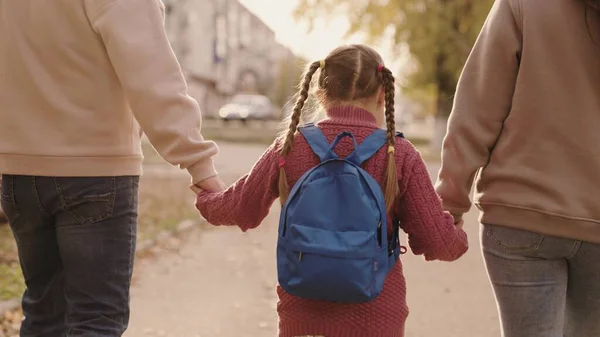 Madre y padre sostienen a una colegiala con una mochila de la mano, familia feliz, caminan por el patio de la escuela en otoño, mamá, papá y niño con la bolsa sobre sus hombros, acompañan al niño al primer grado — Foto de Stock