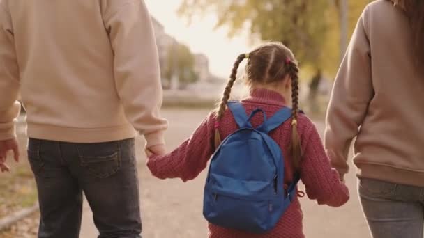Мать и отец держать маленькую школьницу с рюкзаком за руку, счастливая семья, ходить по школьному двору осенью, мама, папа и ребенок с сумкой на плечах, сопровождать ребенка в первом классе — стоковое видео