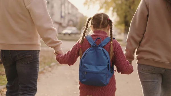 Matka i ojciec trzymają małą uczennicę z plecakiem za rękę, szczęśliwą rodzinę, chodzą jesienią po szkolnym podwórku, mama, tata i dziecko z torbą na ramionach, towarzyszą dziecku do pierwszej klasy — Zdjęcie stockowe