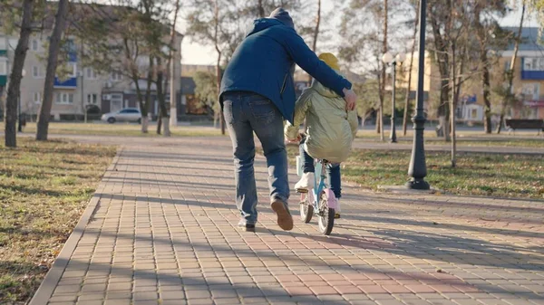Otec učí malé dítě jezdit na kole na silnici v městském parku, dítě točí pedály a kola, šťastná rodina, táta pomáhá dívce jezdit na dceři, hrát v podzimním parku, rodiče a dítě baví společně — Stock fotografie