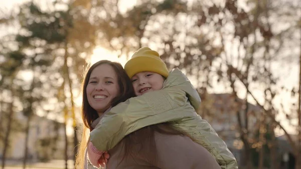 A cheerful mother circles her little daughter in her arms in the glare of city sunset, happy family, life of motherhood, child with her mother laughs hugging, kid with parent in sunlight, walk park — Stock Photo, Image