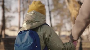 Anne, güz döneminde okul bahçesinde sırt çantalı neşeli bir çocuğu okula götürüyor, okul çantalı küçük bir çocuk annesinin elini tutuyor, mutlu bir aile, ilkokul öncesi eğitim, birinci sınıf öğrencisinin çocukları.