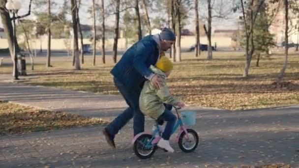 Весёлый отец учит маленького ребенка ездить на двухколесном велосипеде счастливая семья, ребенок мечты привод велосипеда папа играть весело городской парк, ребенок крутит педали колеса, родители занимаются верховой ездой с дочерью — стоковое видео