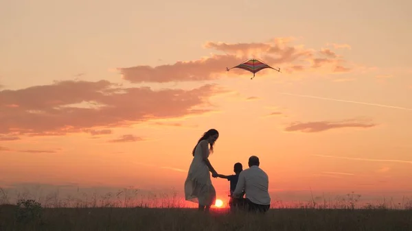 Familia feliz, niño pequeño con padre y madre lanzan cometa voladora en el cielo del atardecer, silueta de mamá, papá y niño juntos, juguete volador vuela alto en su cabeza, sueños de fantasía de los niños, diversión de viaje — Foto de Stock