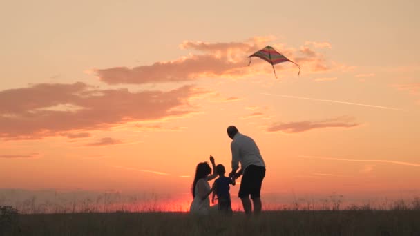 Χαρούμενο παιδί εγκαινιάζει χαρταετό με τους γονείς στο ηλιοβασίλεμα, μικρό παιδί παίζει με τον πατέρα και τη μητέρα υπό το φως του ήλιου, αγόρι γιος με τη μαμά και τον μπαμπά ευτυχισμένη οικογένεια, χαρούμενη ζωή Σαββατοκύριακο στη φύση υπαίθρια ταξίδια — Αρχείο Βίντεο