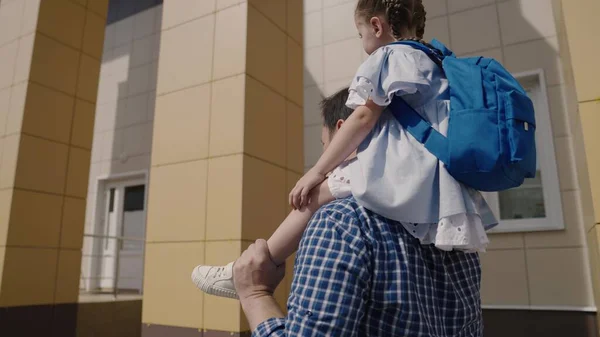 Tata nosi na ramionach małe dziecko z plecakiem do szkoły, dziewczyna z torbą szkolną jeździ swoim ojcem po podwórku szkolnym, szczęśliwa rodzina, szkoła podstawowa, pierwszoklasista z rodzicem chodzi do klasy — Zdjęcie stockowe