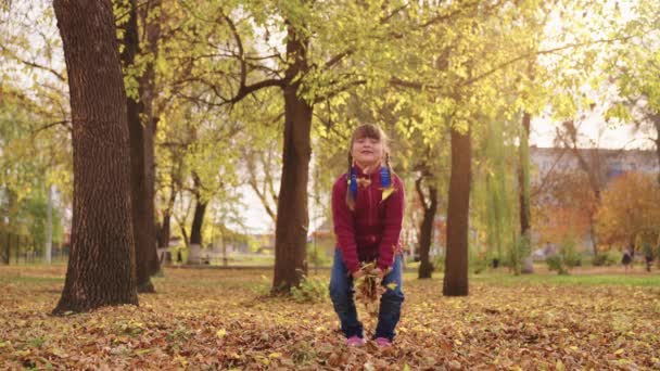 Маленькая девочка с рюкзаком бросает осенние листья и смеется, веселый ребенок играет с лист осенью город-стрит, радостный ребенок весело парк после школьных уроков, счастливая семья, детские фантазии мечты — стоковое видео