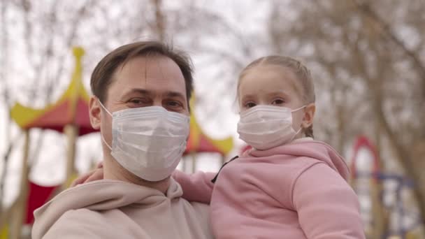 Litet barn i famnen på sin far skyddande medicinsk mask, lycklig barndom, pandemisk coronavirus infektion covid-19, pappa med barn hans armar ler på lekplatsen, flicka dotter stadspark — Stockvideo