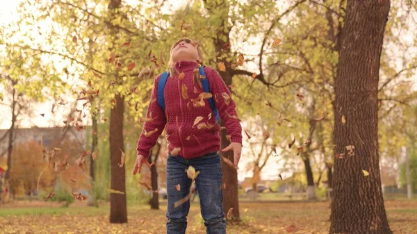 バックパック付きの小さな幸せな女の子は、高い葉を投げ、陽気に笑い、秋の公園で子供の遊び、街の公園で子供の散歩、黄金の葉の秋、秋の季節を見て彼女の顔を上げる — ストック写真