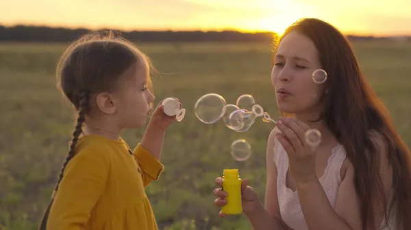 mother with small child blowing round soap bubbles sunset, happy family, cheerful kid and mom are playing with soap foam rays sunlight, girl with parent laughing while playing game, childhood dream