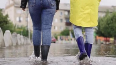 Lastik çizmeli kızlar bir su birikintisinde yürüyor, bir şehir caddesinde sel, yollarda ve asfalt yollarda sel baskını, ıslak çamur birikintisinden farklı yönlere sıçrıyor,