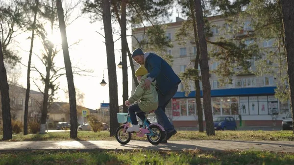 Батько вчить маленьку дитину їздити на велосипеді по дорозі в міському парку, дитина крутить педалі і колеса, щаслива сім'я, тато допомагає дівчинці їздити донькою, грати в осінньому парку, батьки і дитина розважаються разом — стокове фото