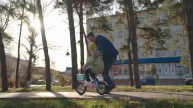 Babası küçük bir çocuğa parkta bisiklet sürmeyi öğretiyor, çocuk pedal çeviriyor ve tekerlek çeviriyor, mutlu aile, baba kızına binmesine yardım ediyor, sonbahar parkında oynuyor, ebeveyn ve bebek birlikte eğleniyor.