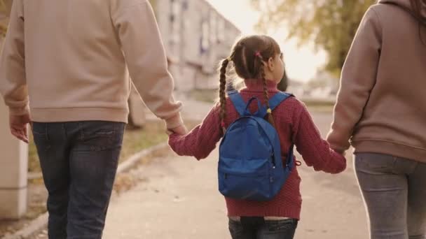 Anya és apa tart egy kis iskolás lány egy hátizsák a kezét, boldog család, séta az iskola udvarán ősszel, anya, apa és a gyerek táskával a vállukon, elkíséri a gyermeket az első osztályban — Stock videók