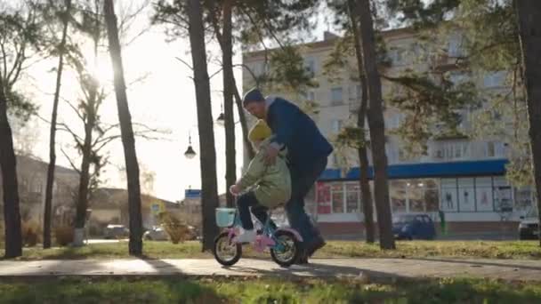 Батько вчить маленьку дитину їздити на велосипеді по дорозі в міському парку, дитина крутить педалі і колеса, щаслива сім'я, тато допомагає дівчинці їздити донькою, грати в осінньому парку, батьки і дитина розважаються разом — стокове відео
