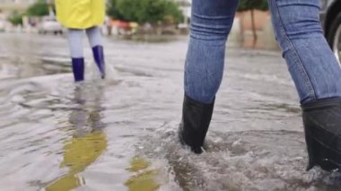 Yağmurluklu ve lastik çizmeli kızlar yağmur altında yol boyunca yürür, ayakları su birikintileri şehrinde yürür, kenarlara su sıçratır, sel caddede, araba su üstünde gider.