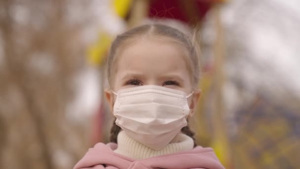 Niña se pone una máscara médica en la cara, protege al niño de la infección por coronavirus, brote de pandemia covid 19, niño camina en el patio de recreo, cuidar de la respiración del bebé, la infancia en modo máscara — Vídeos de Stock