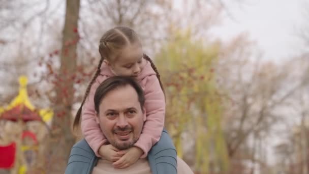 Дитина сидить на плечах бородатого батька і сміється, щаслива сім'я, дитина посміхається обіймаючи тата, веселий батько зі своєю маленькою улюбленою дитиною, дівчинка дочка гуляє на відкритому повітрі природа міського парку — стокове відео