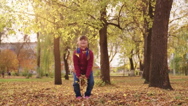 Маленькая девочка с рюкзаком бросает осенние листья и смеется, веселый ребенок играет с лист осенью город-стрит, радостный ребенок весело парк после школьных уроков, счастливая семья, детские фантазии мечты — стоковое видео