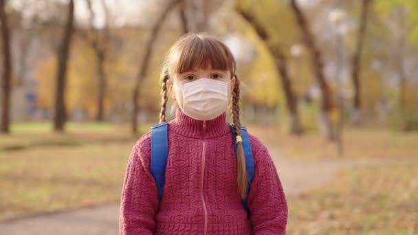 Dziewczynka z plecakiem stoi w masce medycznej na twarzy, pandemia koronawirusa, covid-19, aby pokonać wirusa i zachować zdrowie dzieci, dzieciak z torbą szkolną ramiona, park dziecka iautumn — Wideo stockowe