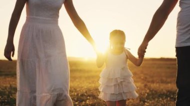 Mutlu bebek gün batımında anne, baba ve çocukla birlikte gülümsüyor. Güneşte yürüyorlar. Çocuklarını elleriyle tutuyorlar, küçük kızlarına bakıyorlar.