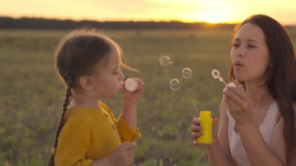 Madre con un niño pequeño que sopla burbujas de jabón redondas puesta de sol, familia feliz, niño alegre y mamá están jugando con los rayos de espuma de jabón la luz del sol, niña con los padres riendo mientras juegan juego, sueño de la infancia — Vídeo de stock