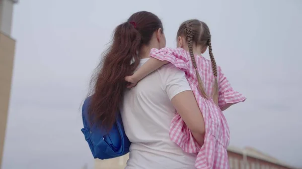 Mor bär en liten dotter med en ryggsäck i famnen till skolan, lära ett barn i grundskolan, en lycklig familj, se ett barn med en skolväska, få kunskap om det framtida livet — Stockfoto