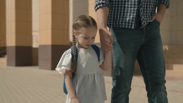 Тато везе маленьку дочку до школи, дівчинка з рюкзаком тримає свого батька за руку, система освіти для дітей, щаслива сім'я, весела дитина з батьками йде через — стокове відео