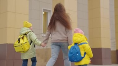 Anne, sırt çantalı çocukları alıp okula götürür, ilkokul öğrencileri sınıfa koşar, anne ve kız çocukları birinci sınıfa gider, okul çantası sırtında olur.