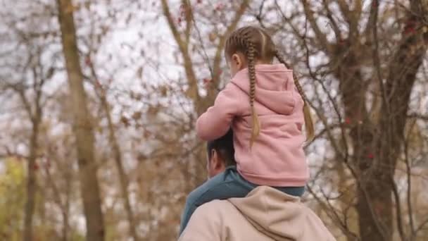 Çocuk babasının boynuna oturur, baba çocuğu sonbahar parkında omuzlarında taşır, mutlu bir aile, bebek gezintisi sevgili ebeveyn, büyüleyici küçük kız açık havada gezer, gökyüzünün arka planına doğru yol alır. — Stok video
