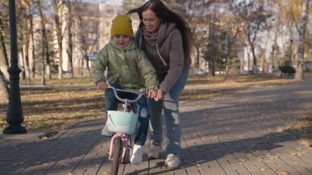 Mother teaches little child to ride bike in the city, kid rides on sidewalk, happy family, baby plays and laughs with mom, learn ride, make childhood dream come true girls, daughter and mom are funny — Stock Video