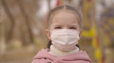 Küçük kız tıbbi maske takarak gülümsüyor, zaman koronavirüsü salgını, sağlıklı vücudu viral enfeksiyondan koruyor covid-19, çocuk parkı, çocuk yaşam tarzı, halka açık yerlerde olma kuralları.