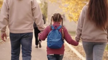 Sırt çantalı küçük bir çocuk anne ve babasını elleriyle tutuyor, anne ve babasıyla okul bahçesinde geziyor, mutlu bir aile, ilk aramada acele ediyor, ilkokul, okul çantası bebek omuzlarında.