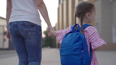 Sırt çantalı küçük bir kız annesinin elinden tutup okula gidiyor. Annesiyle birlikte okul bahçesinde okula gidiyor. Okul çantalı bir çocuk ilk derse yetişmek için acele ediyor.