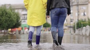 Lastik çizmeli kızlar bir su birikintisinde yürüyor, bir şehir caddesinde sel, yollarda ve asfalt yollarda sel baskını, ıslak çamur birikintisinden farklı yönlere sıçrıyor,