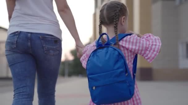 Маленькая девочка с рюкзаком держит свою мать за руку и идет в школу, ребенок с мамой ходить в школу в школьном дворе, ребенок со школьной сумкой спешит на первый урок, подготовительное дошкольное воспитание — стоковое видео