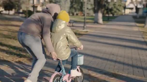 Vidám gyermek tanul lovagolni kétkerekű kerékpár az anyjával, boldog család, gyerek nevet pedál kerék vezetés közben, baby play city park az anyjával, szülő segít a lánynak tanulni előre — Stock videók
