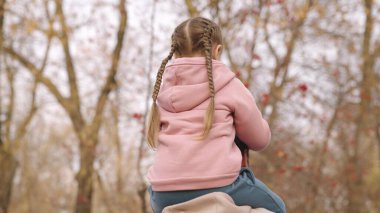 Çocuk babasının boynuna oturur, baba çocuğu sonbahar parkında omuzlarında taşır, mutlu bir aile, bebek gezintisi sevgili ebeveyn, büyüleyici küçük kız açık havada gezer, gökyüzünün arka planına doğru yol alır.