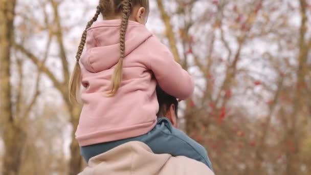 子供は父親の首に座り父親は秋の公園で子供を肩に担いで幸せな家族赤ちゃんは最愛の親に乗る魅力的な少女は空の背景を背景にオープンエアの性質に乗る — ストック動画