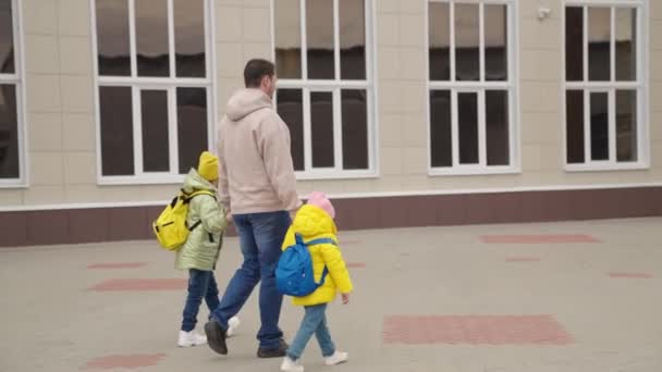Otec vede malé děti s batohy, které se drží za ruce do školy, dětská školka, táta a dcery spěchají do třídy, šťastná rodina, táta vzal dítě po školním dni, předškolní výchova — Stock video