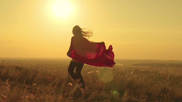 Happy teenage girl runs playing superhero at sunset, child on a green field in a red raincoat toy in the rays of light, childrens dream game, kid dreams of becoming a superhero in glare of the sun.