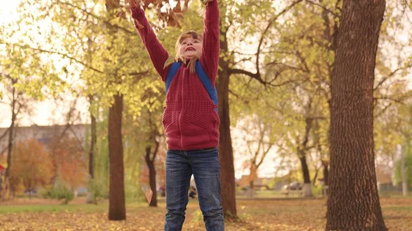 Klein gelukkig meisje met een rugzak gooit bladeren hoog en lacht vrolijk, kind spelen in de herfst park, kind wandelingen in het stadspark, verhogen haar gezicht kijken naar de gouden blad vallen, herfst seizoen — Stockfoto