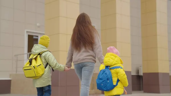 Madre sostiene a los niños con mochilas a mano y los lleva a la escuela, los estudiantes de la escuela primaria se apresuran a la clase, la madre y las hijas van al primer grado, bolsa de la escuela en sus hombros detrás de su espalda — Foto de Stock